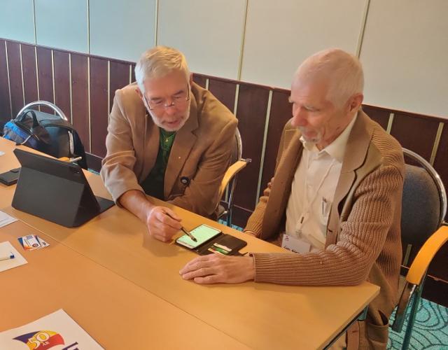 två personer sitter vid ett bord och ser på en smarttelefon, den ena pekar på telefonen